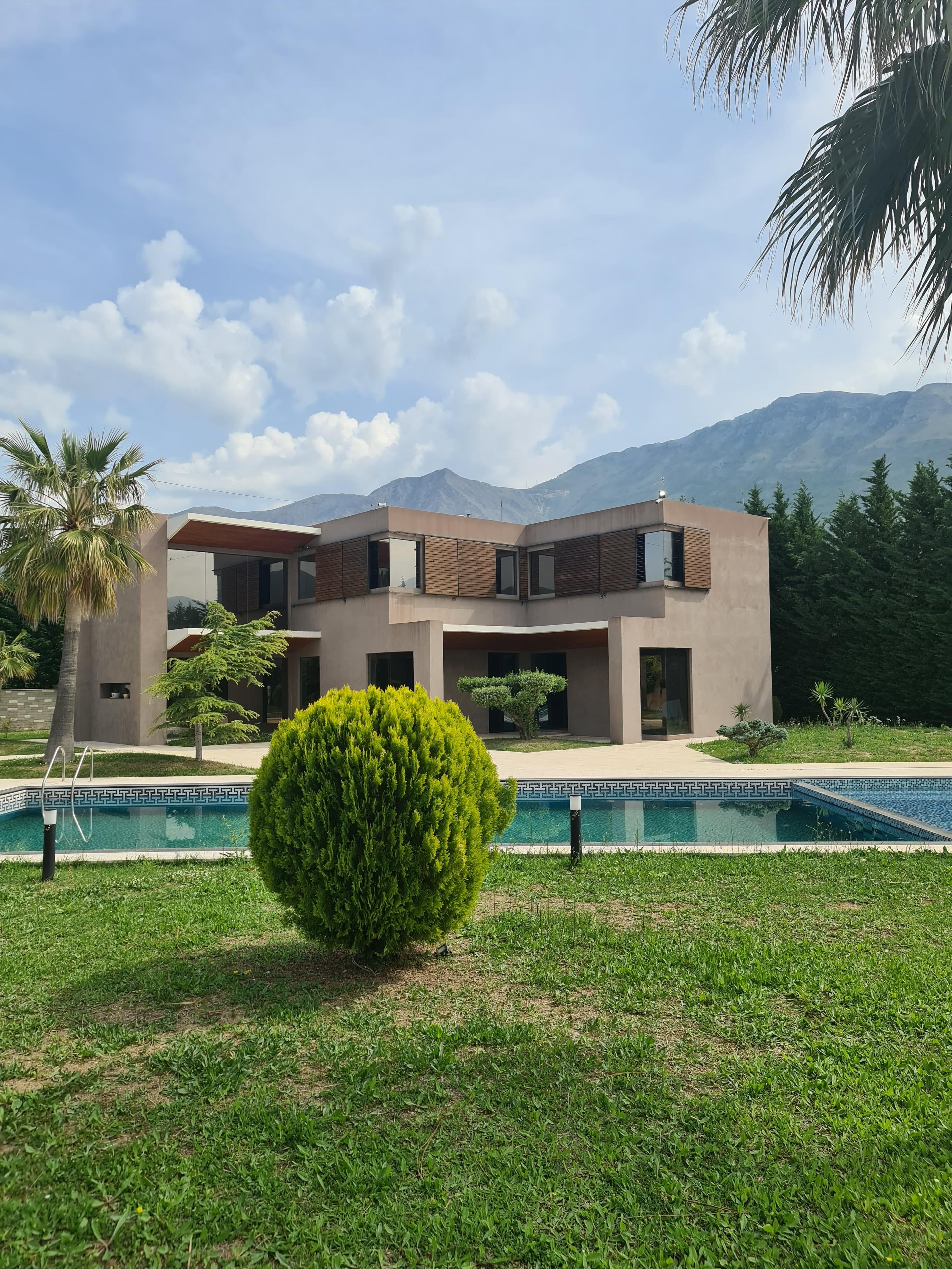 Casa moderna in vendita a Vlore, Albania, con piscina privata, circondata da un giardino curato e palme. Sullo sfondo sono visibili montagne pittoresche e cielo azzurro con nuvole.