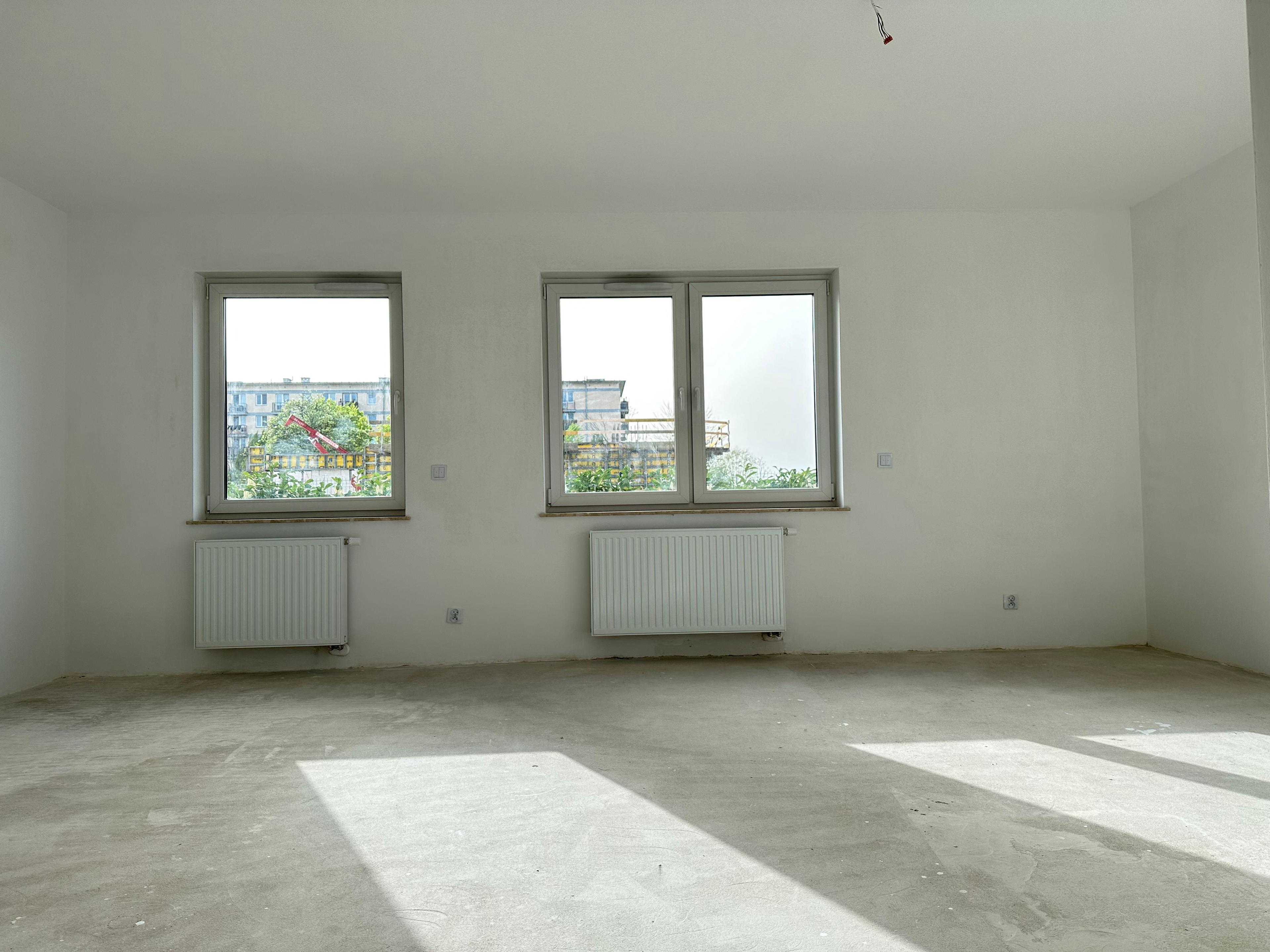Salon spacieux dans un nouvel appartement en état de développement – trois grandes fenêtres offrant une lumière naturelle, murs blancs et sol brut permettant un aménagement libre. Vue sur la ville et radiateurs modernes sous les fenêtres.