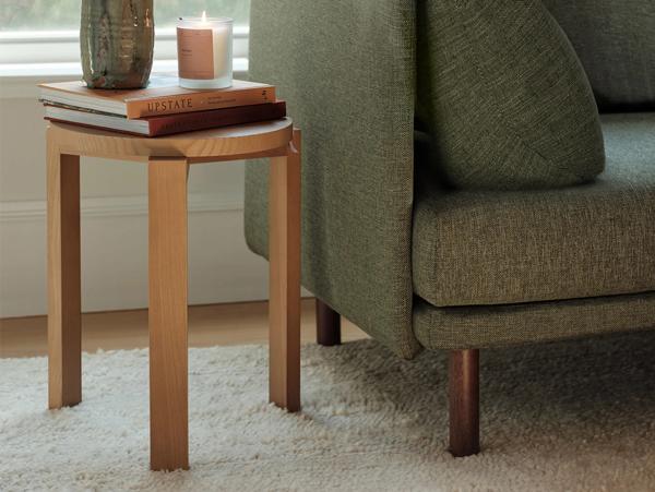 Round wooden side table with stacked books and lit candle next to olive green upholstered chair on white rug
