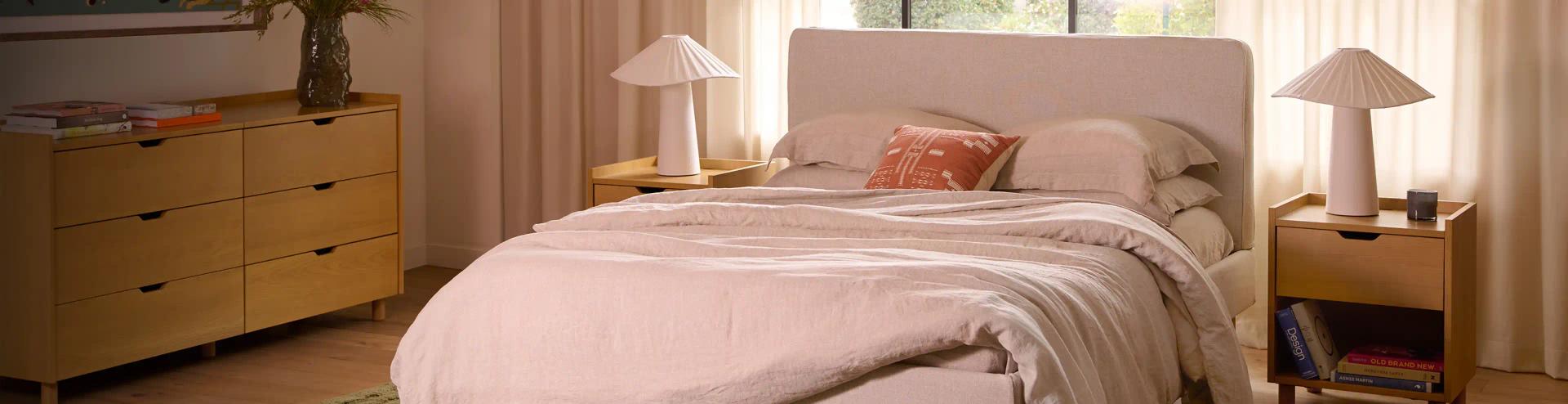 Hero Image Cozy bedroom featuring a soft bed with neutral linens, stylish lamps, and a wooden dresser adorned with books and decorative items.
