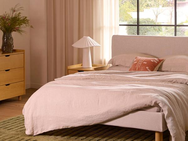 Pink upholstered bed with linen bedding, wooden nightstand, and pleated table lamp in warm-toned bedroom