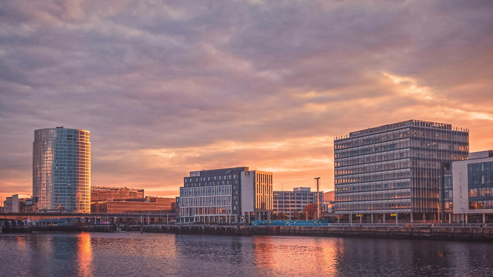 Belfast waterfront at sunset, with the River Lagan reflecting orange light and the Obel Tower on the left of the skyline.