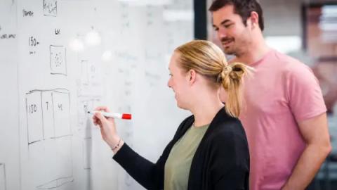 A woman sketching a wireframe layout on a whiteboard while a colleague looks on.