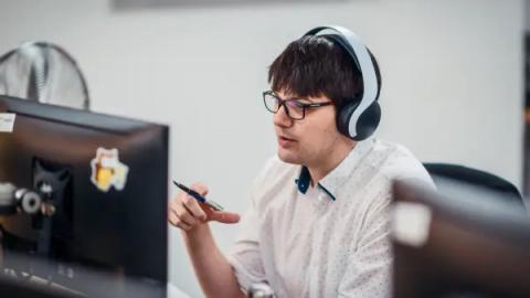 A man wearing headphones talking during a video call at his desk, holding a pen