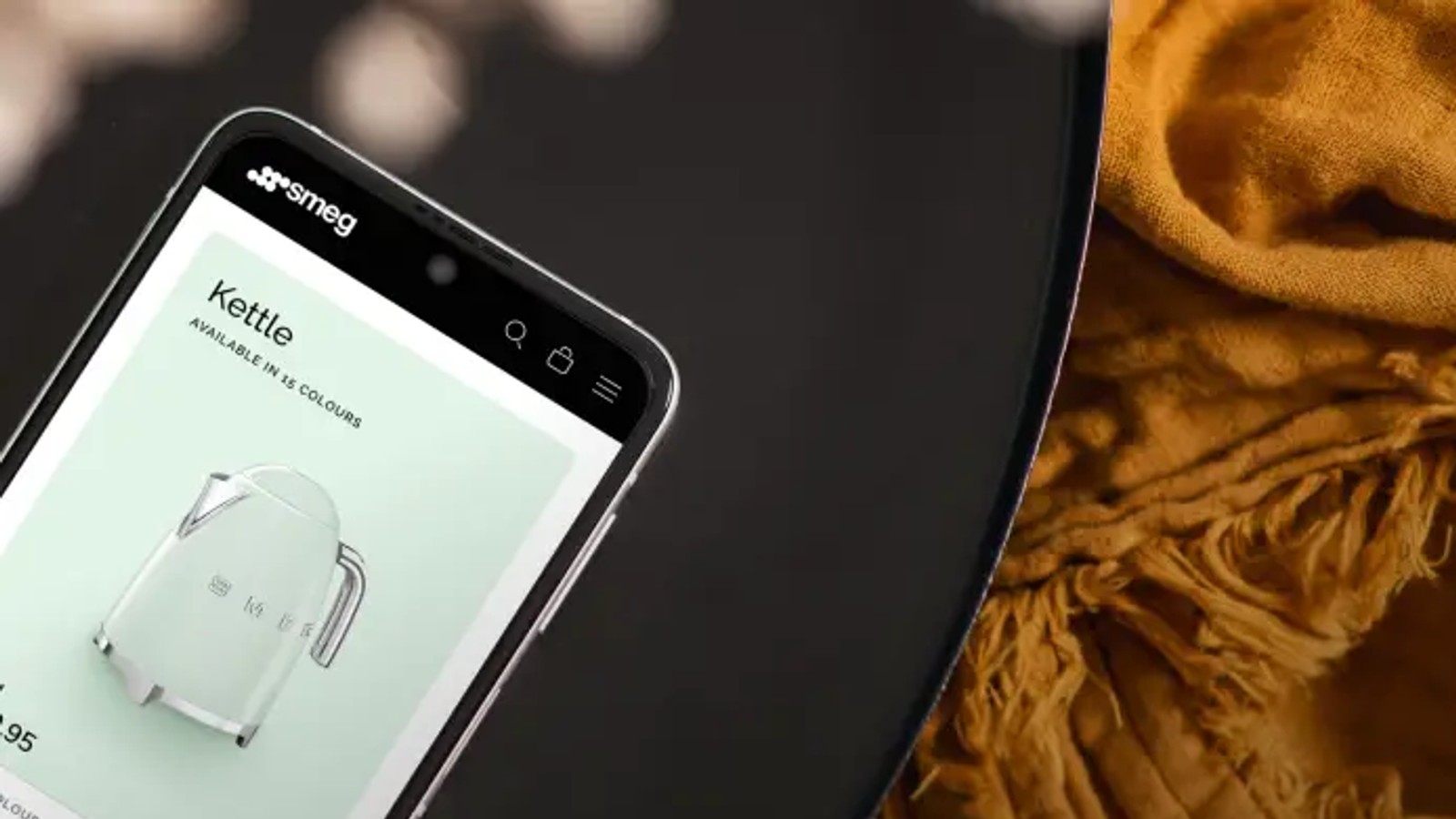 A smartphone displaying the Smeg website's kettle product page rests on a dark round table, next to a folded mustard-yellow textile. Text overlay reads: What is Optimizely?