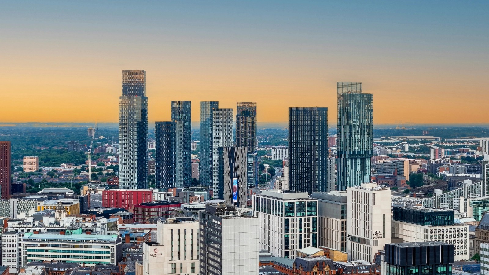 Aerial view of the Manchester skyline at sunset, with the Deansgate Square towers and Beetham Tower against an orange sky.