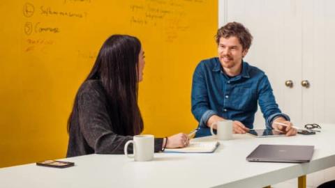 Two colleagues talking across a table in a meeting room, with a yellow whiteboard of handwritten notes behind them.