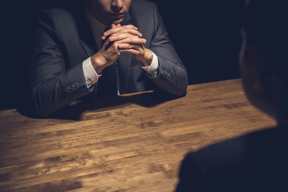 Image A man sitting alone at a table with his hands clasped in a dim room