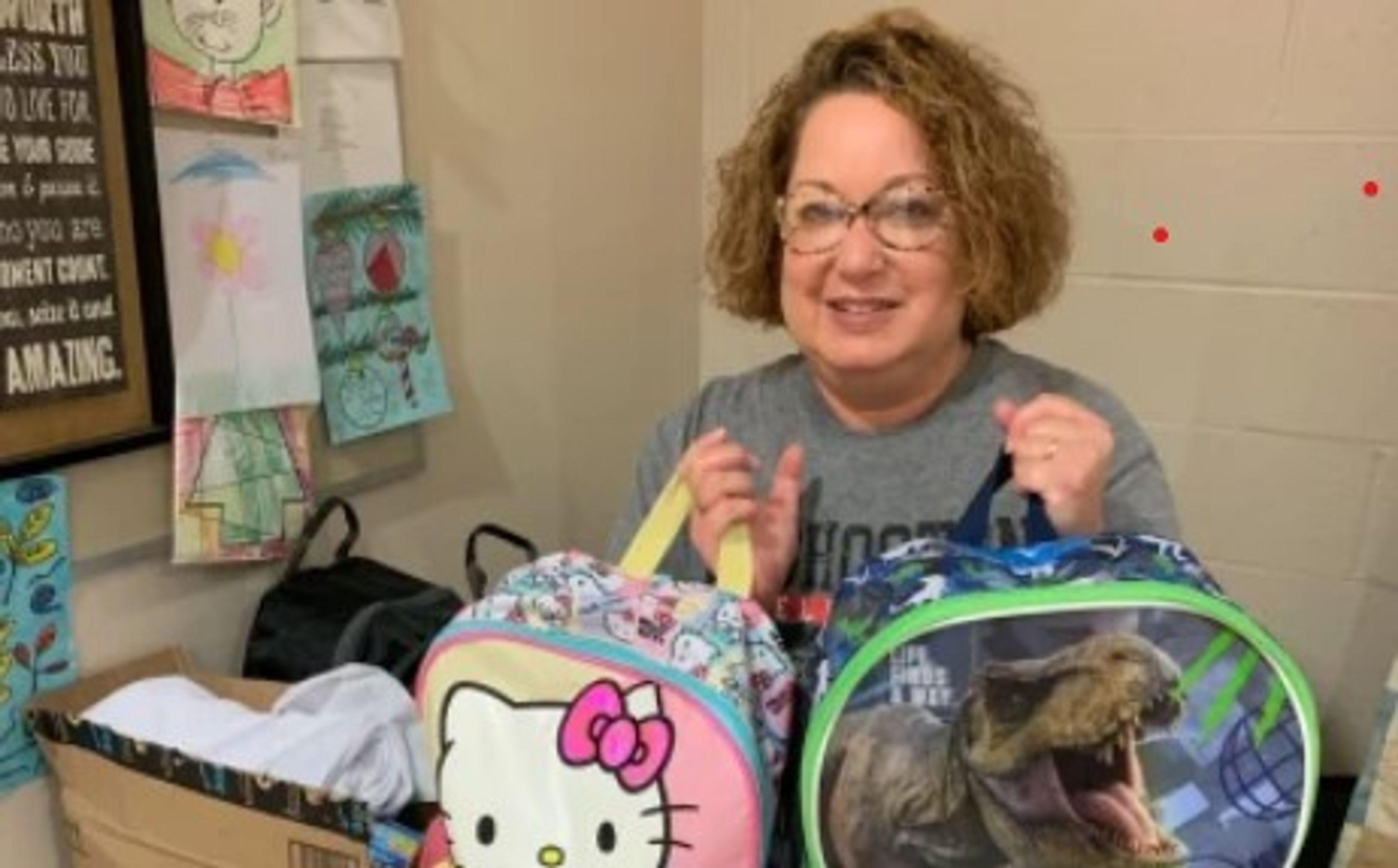 A woman with curly hair smiles while holding two lunchboxes—one with Hello Kitty and one with a dinosaur. Children’s artwork is displayed on the wall behind her.