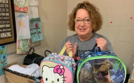 A woman with curly hair smiles while holding two lunchboxes—one with Hello Kitty and one with a dinosaur. Children’s artwork is displayed on the wall behind her.