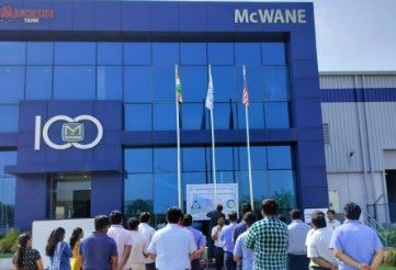 A group of people stands facing three flagpoles outside a blue glass building with “McWANE” signage and a “100” logo, likely attending a flag-raising event or ceremony.