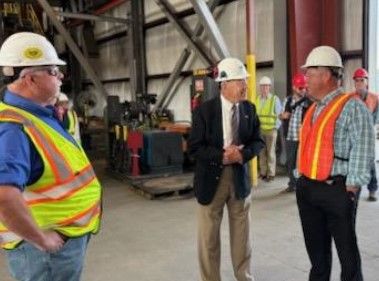 Several men wearing hard hats and safety vests stand and talk inside an industrial building with metal beams and equipment visible in the background.