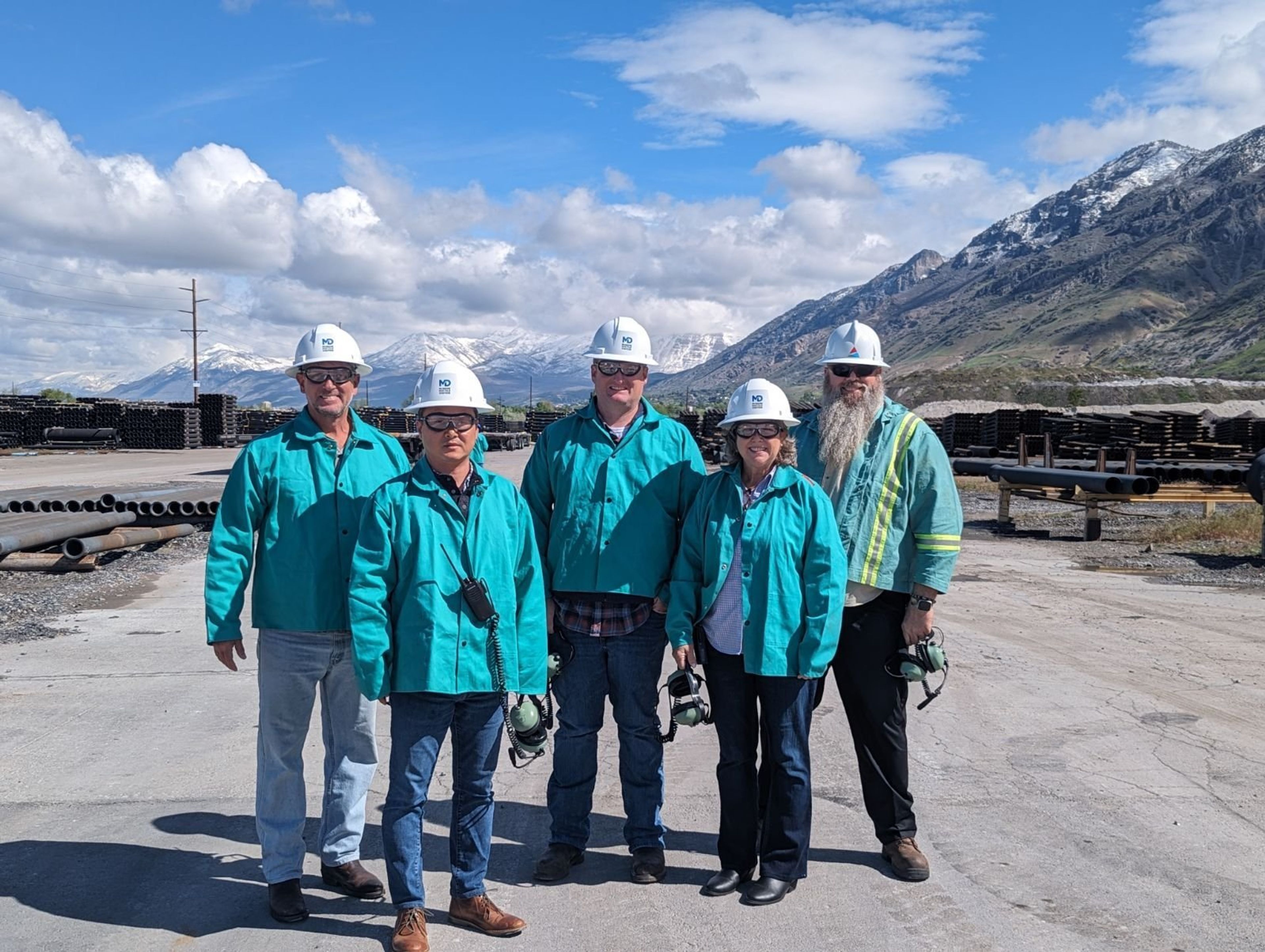 Five people in teal jackets and hard hats stand on a paved site with industrial pipes and mountains in the background, under a partly cloudy sky.