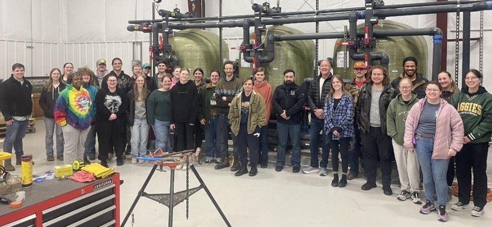 A group of about 30 people poses and smiles together indoors in front of large industrial equipment. Some wear casual clothing, others wear jackets or sweatshirts. The setting looks like a workshop or lab.