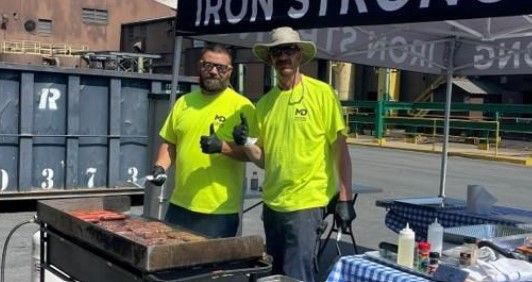 Two men in neon yellow shirts grill meat at an outdoor event under a canopy. One gives a thumbs up. Industrial buildings and a large dumpster are visible in the background.