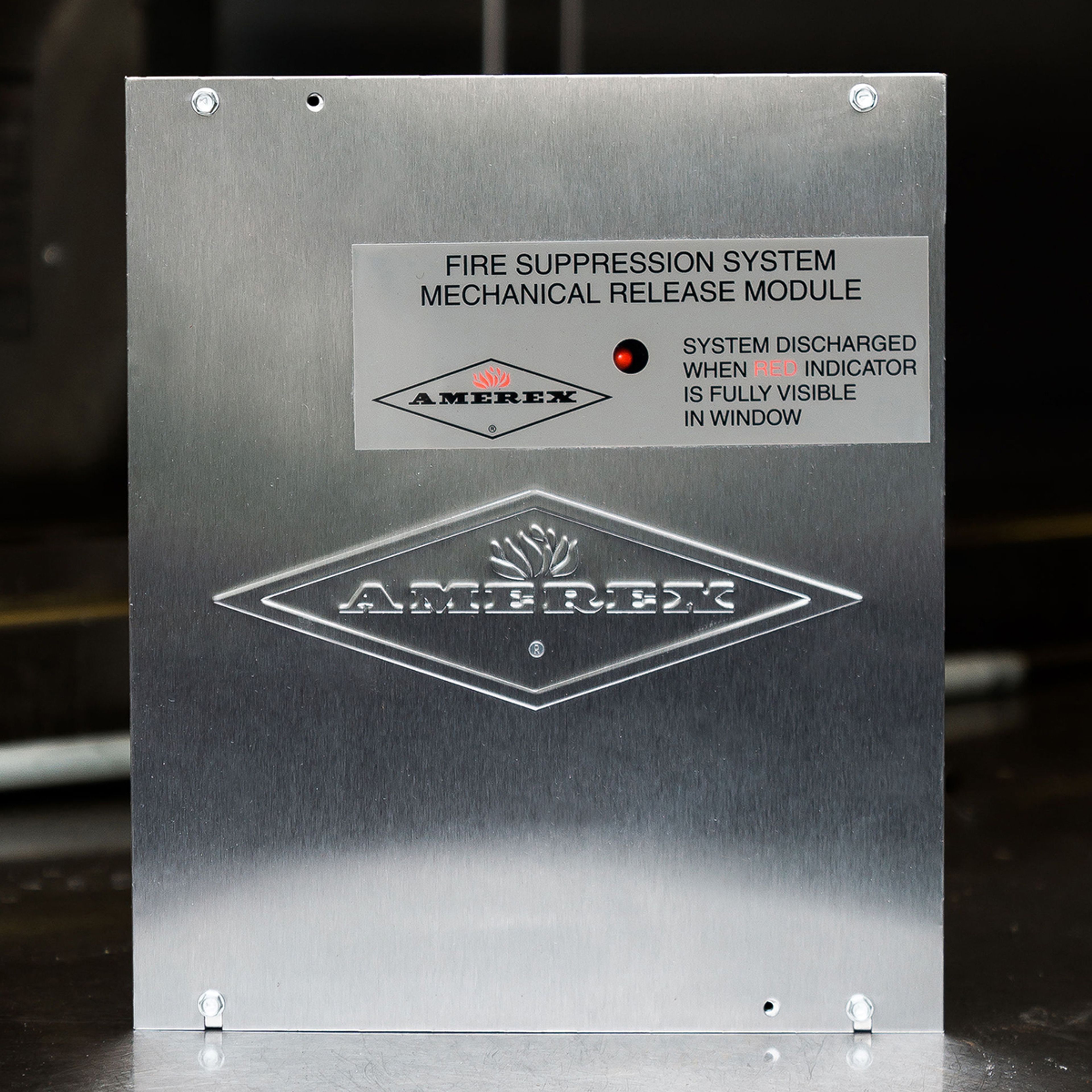 A metal Amerex fire suppression system mechanical release module with a red indicator light and label instructions is shown against a dark background.