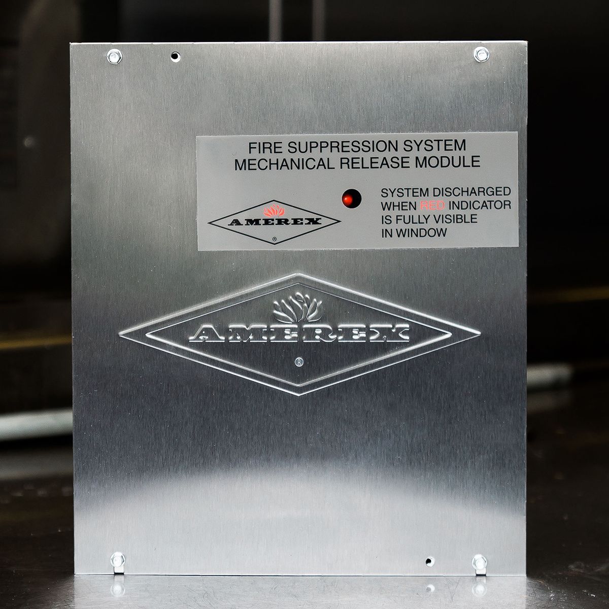 A metal Amerex fire suppression system mechanical release module with a red indicator light and label instructions is shown against a dark background.