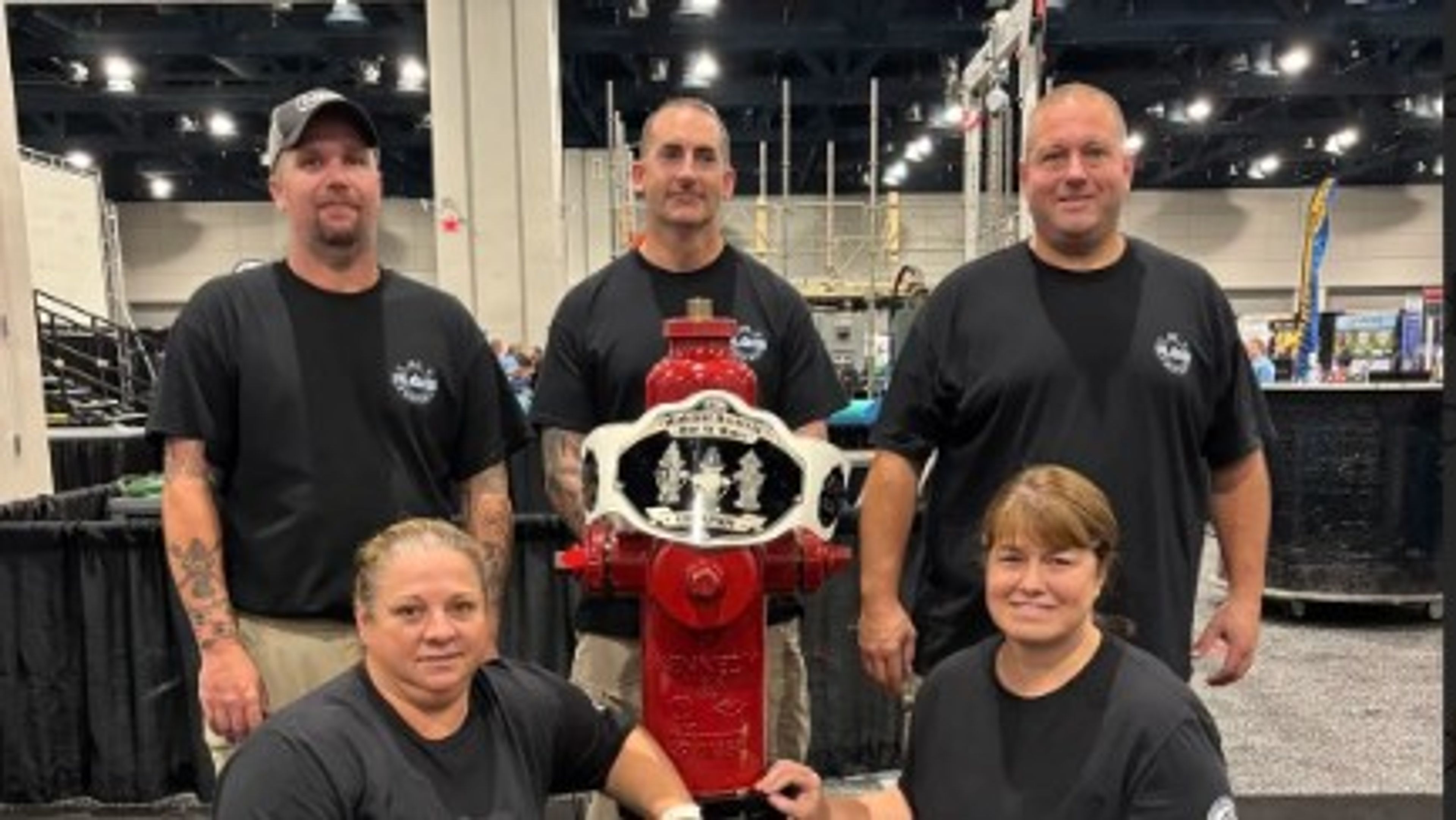Five people in black shirts pose around a red fire hydrant with a championship-style belt. The group stands indoors at an event, with booths and gym equipment visible in the background.