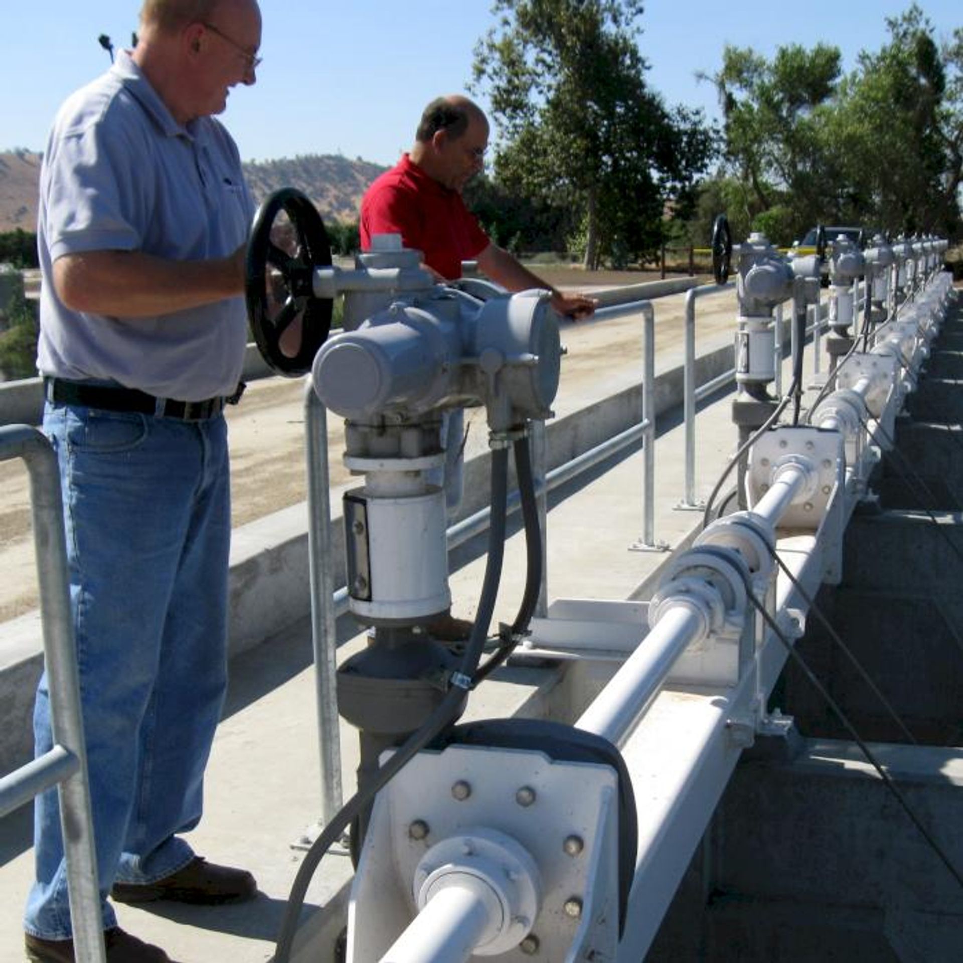 Two men stand on a concrete platform outdoors, operating large valve controls connected to a long pipeline with multiple actuators. Trees and hills are visible in the background.