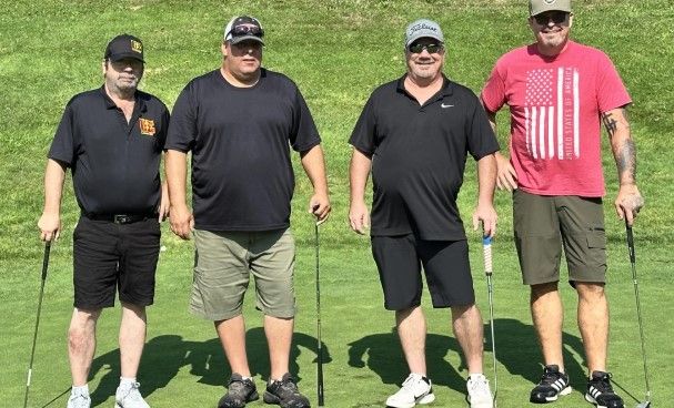 Four men stand on a golf course in casual clothes, each holding a golf club, with green grass in the background.