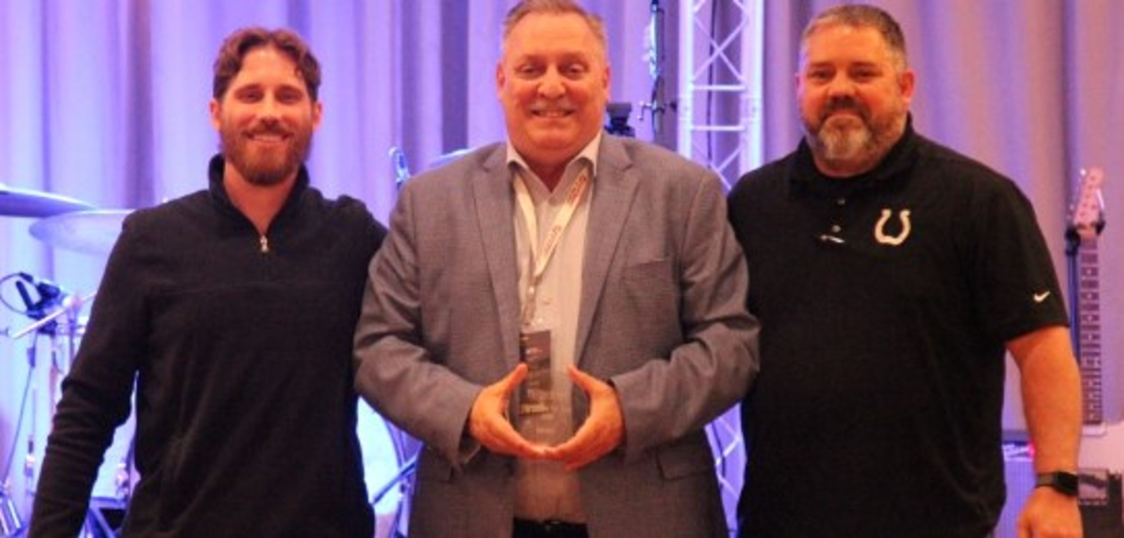 Three men stand together, smiling. The man in the center holds an award. The background shows stage lights and musical instruments, suggesting an event or ceremony.