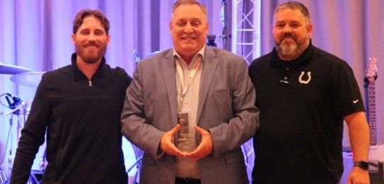Three men stand together, smiling. The man in the center holds an award. The background shows stage lights and musical instruments, suggesting an event or ceremony.