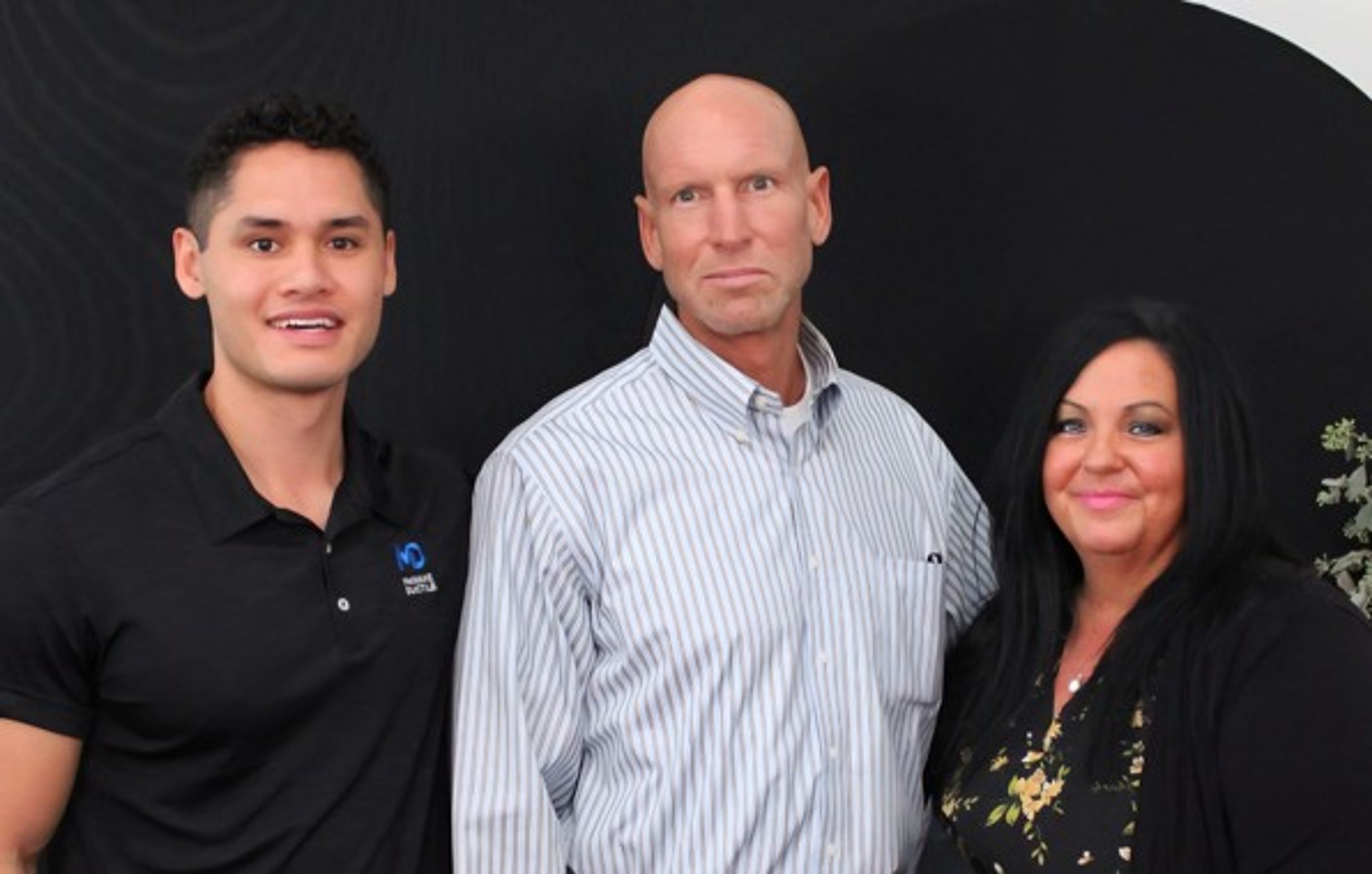 Three people are standing together and smiling in front of a dark background. The man on the left wears a black polo shirt, the man in the middle wears a striped shirt, and the woman on the right has long black hair.