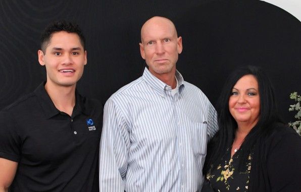 Three people are standing together and smiling in front of a dark background. The man on the left wears a black polo shirt, the man in the middle wears a striped shirt, and the woman on the right has long black hair.