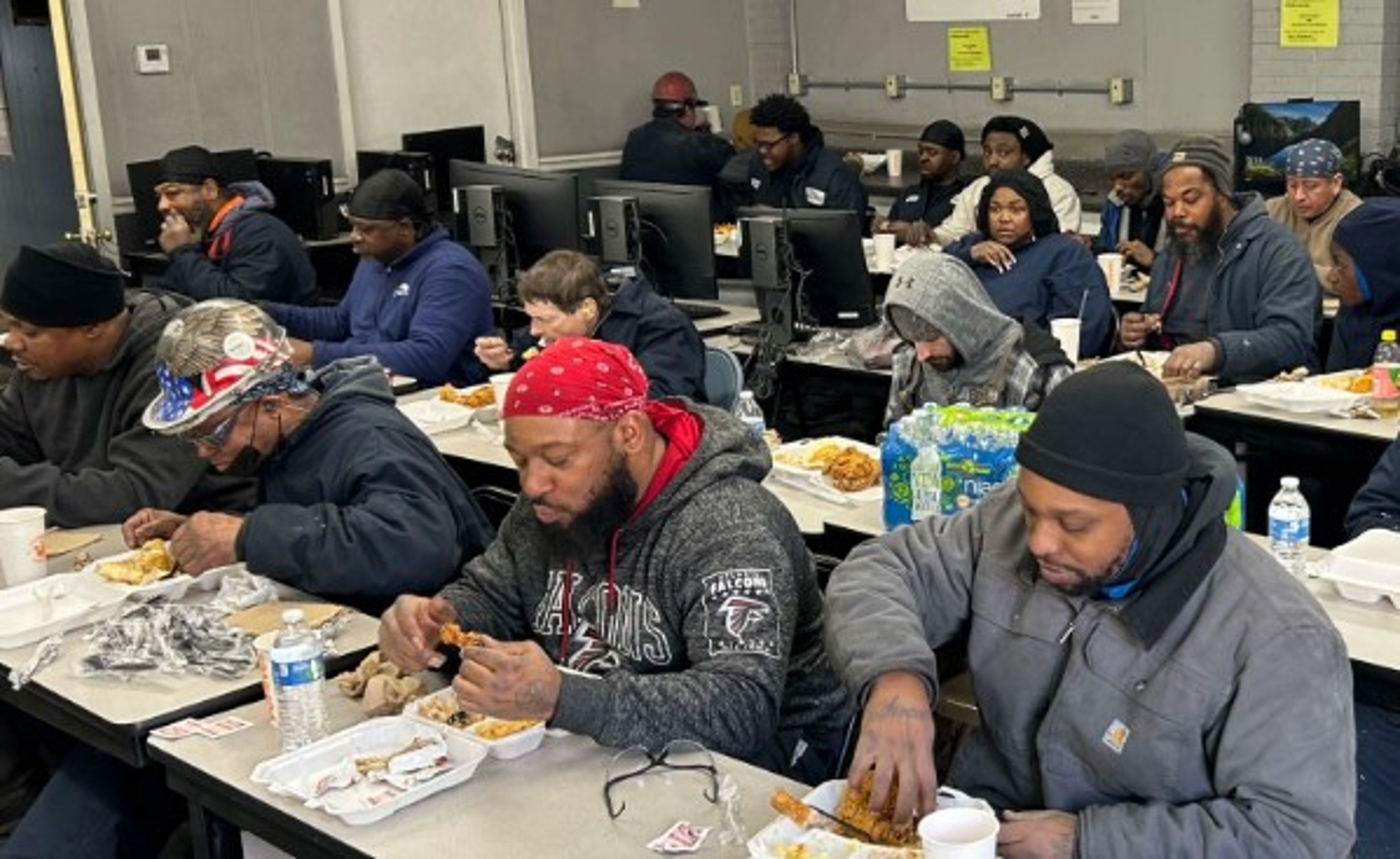 A group of people eat meals together at long tables in a room with computers. Most wear hats or bandanas and winter clothing. Bottled water and boxed lunches are visible on the tables.