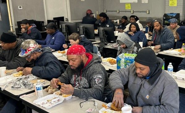 A group of people eat meals together at long tables in a room with computers. Most wear hats or bandanas and winter clothing. Bottled water and boxed lunches are visible on the tables.