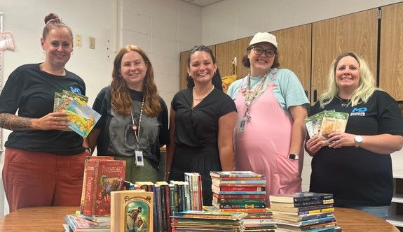 Five women stand behind a table filled with stacks of books in a classroom, smiling at the camera. Some are holding books, and cabinets and white brick walls are visible in the background.