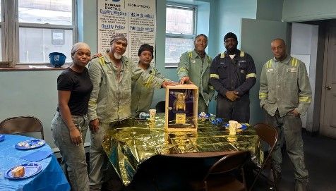 Six people in work uniforms and casual clothes stand around a table with a gold tablecloth and centerpiece in a decorated room, smiling at the camera. Blue plates and drinks are on the table.