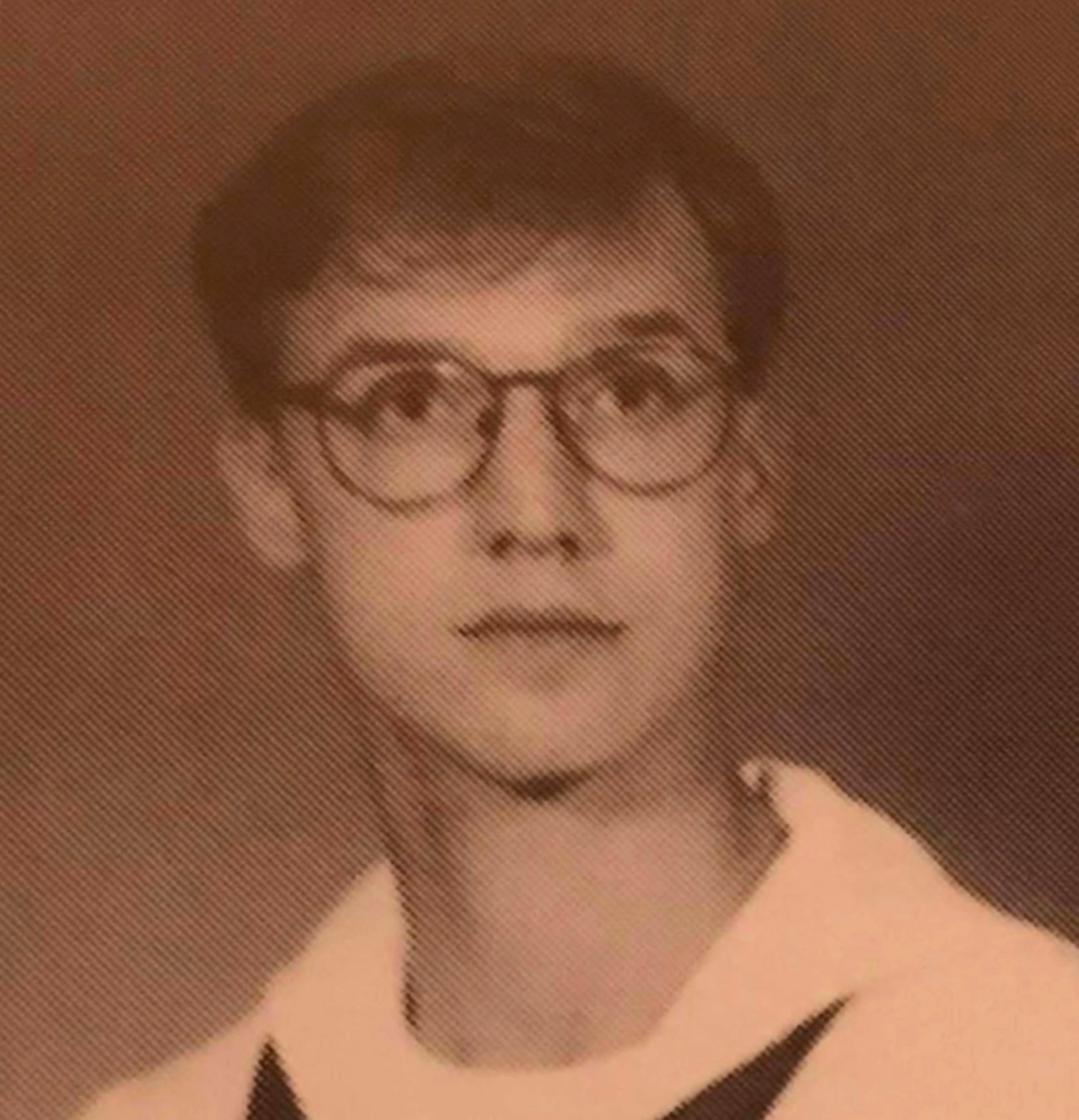 A young person with short hair and glasses wears a light-colored top with a dark collar, looking at the camera with a neutral expression against a plain background. The photo has a sepia tone.