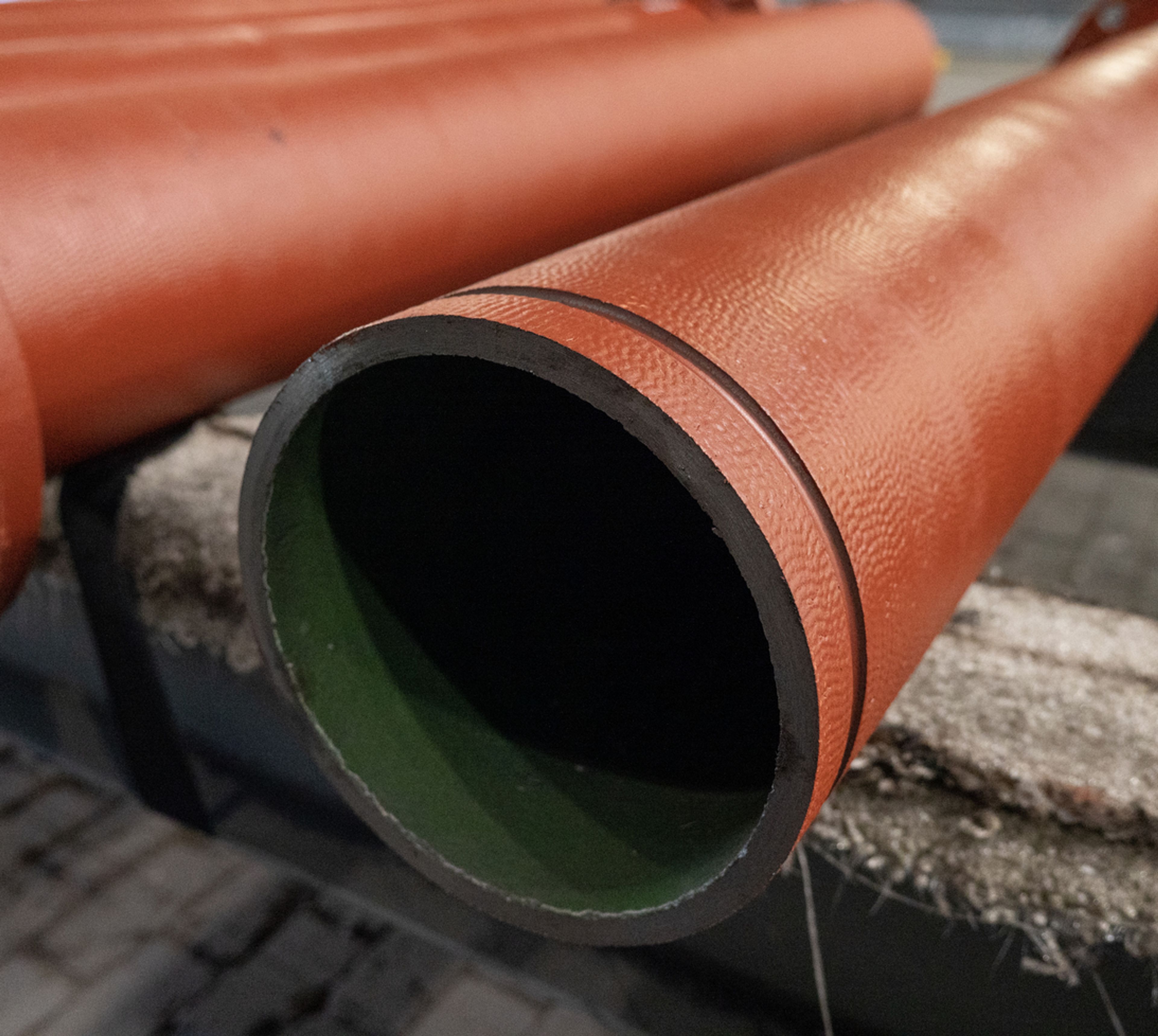 A close-up of a large, cylindrical, red-brown industrial pipe with a green inner lining, resting on a textured surface.