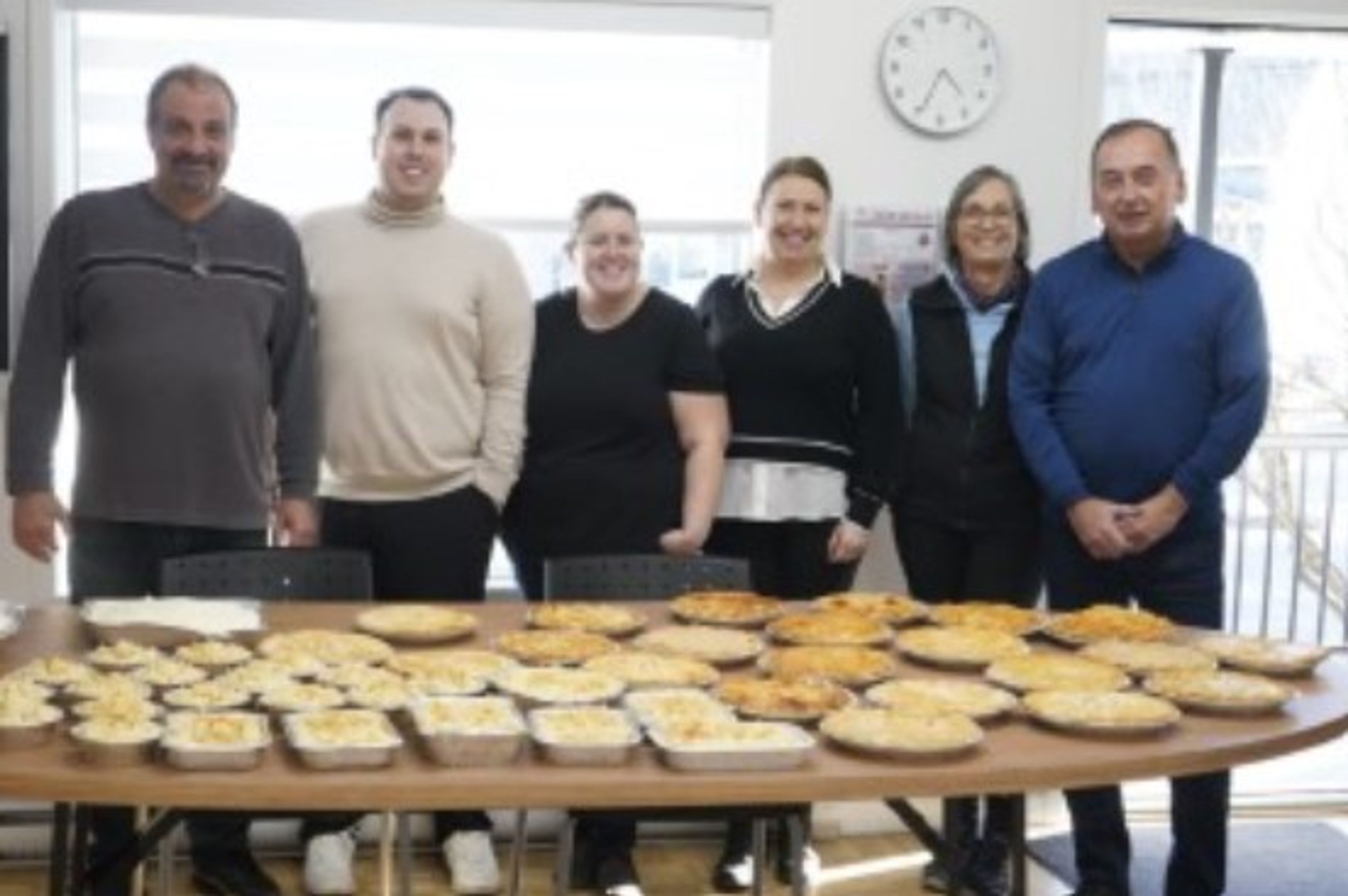 Six people stand behind a table filled with baked pies and casseroles. There is a clock on the wall behind them showing 11:10. Light from large windows brightens the room.