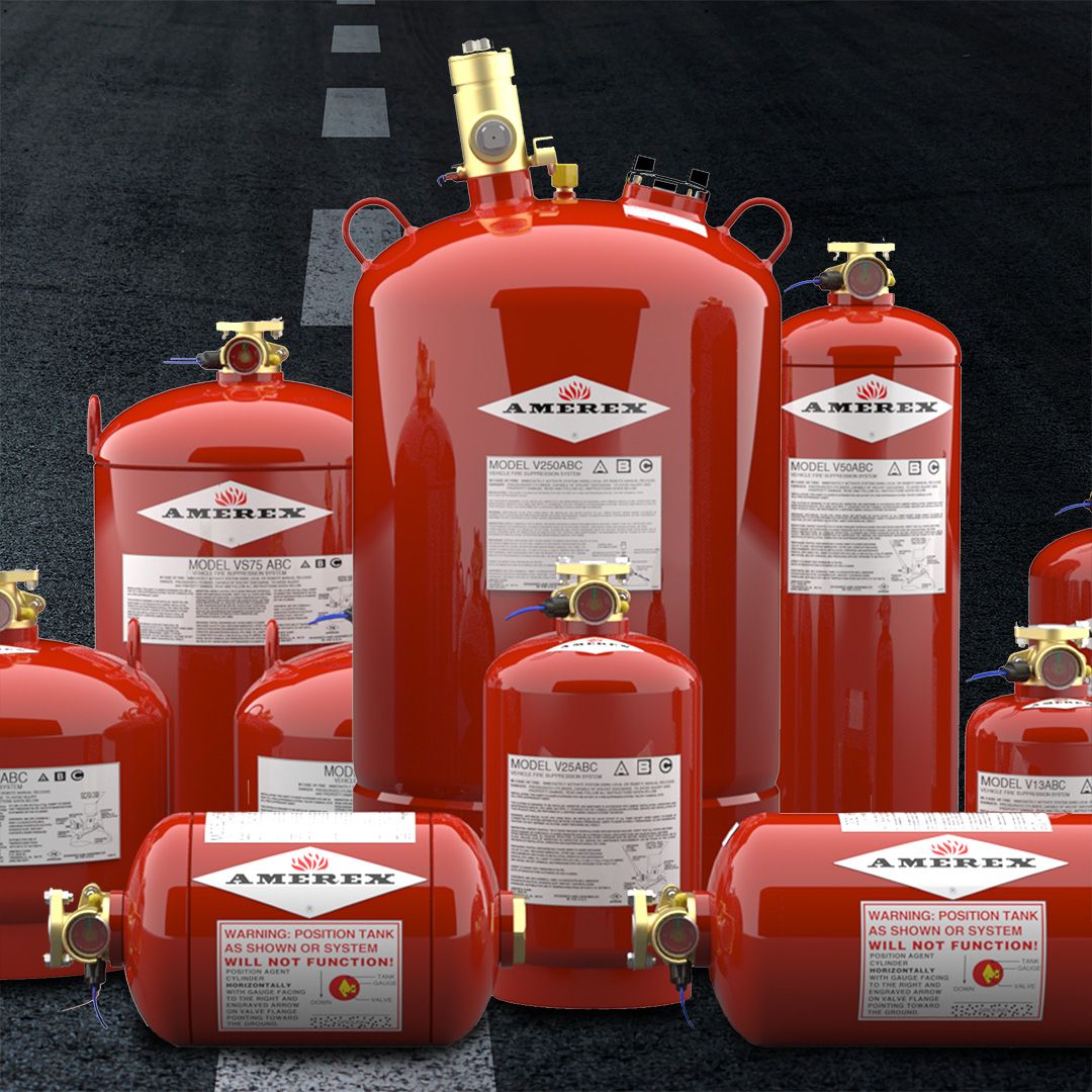 Several red AMEREX fire suppression tanks of various sizes are arranged on an asphalt road, with prominent white labels and brass valves. The labels contain warning and model information.