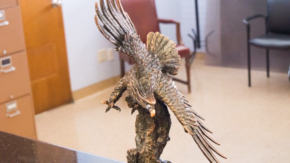 A bronze eagle statue with wings spread, perched on a rock, sits on a desk in an office setting with chairs and cabinets in the background.
