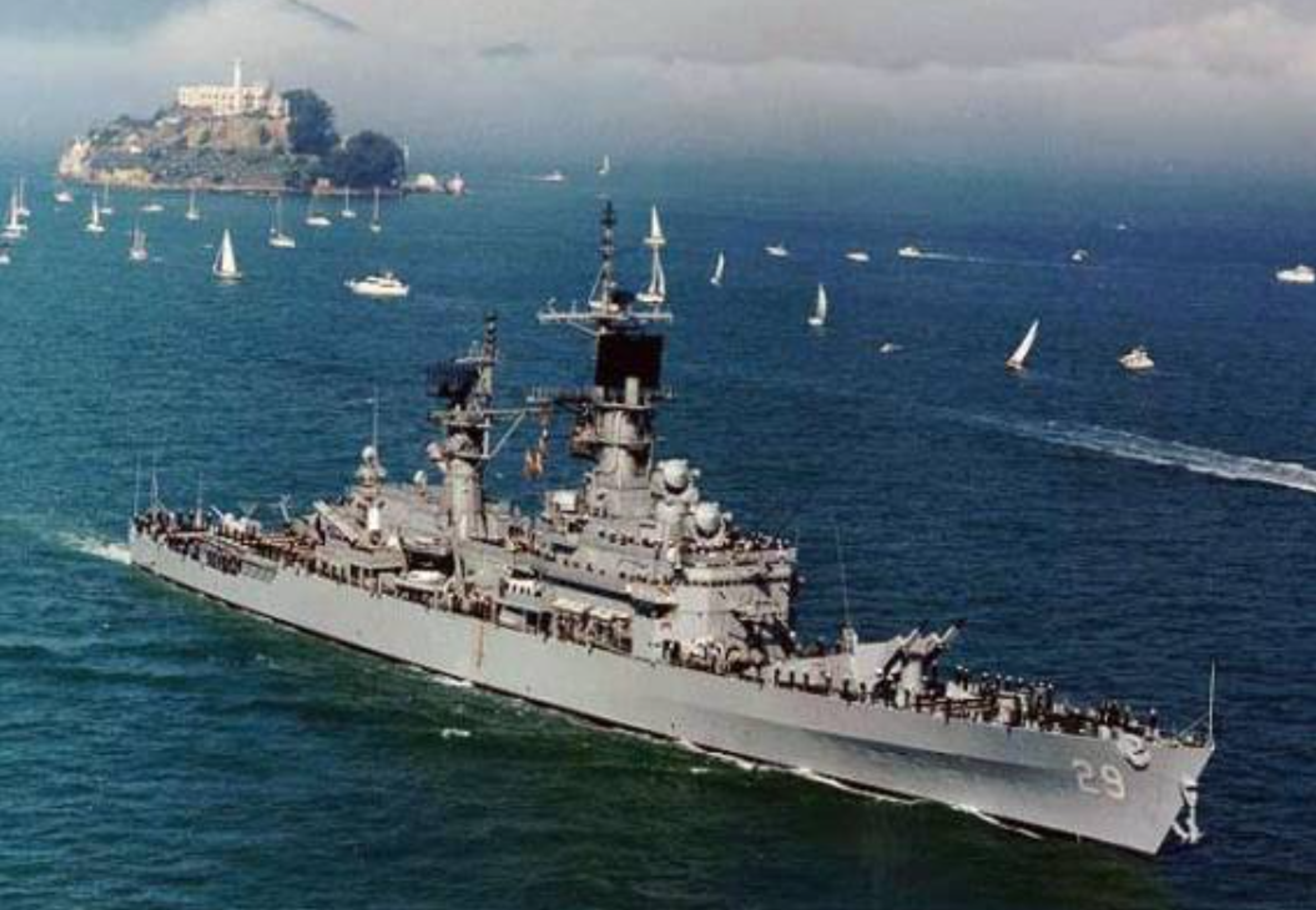 A large gray naval ship sails in blue water with many small sailboats nearby; an island with buildings is visible in the background under a partly cloudy sky.