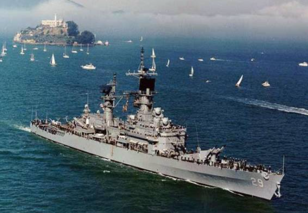 A large gray naval ship sails in blue water with many small sailboats nearby; an island with buildings is visible in the background under a partly cloudy sky.