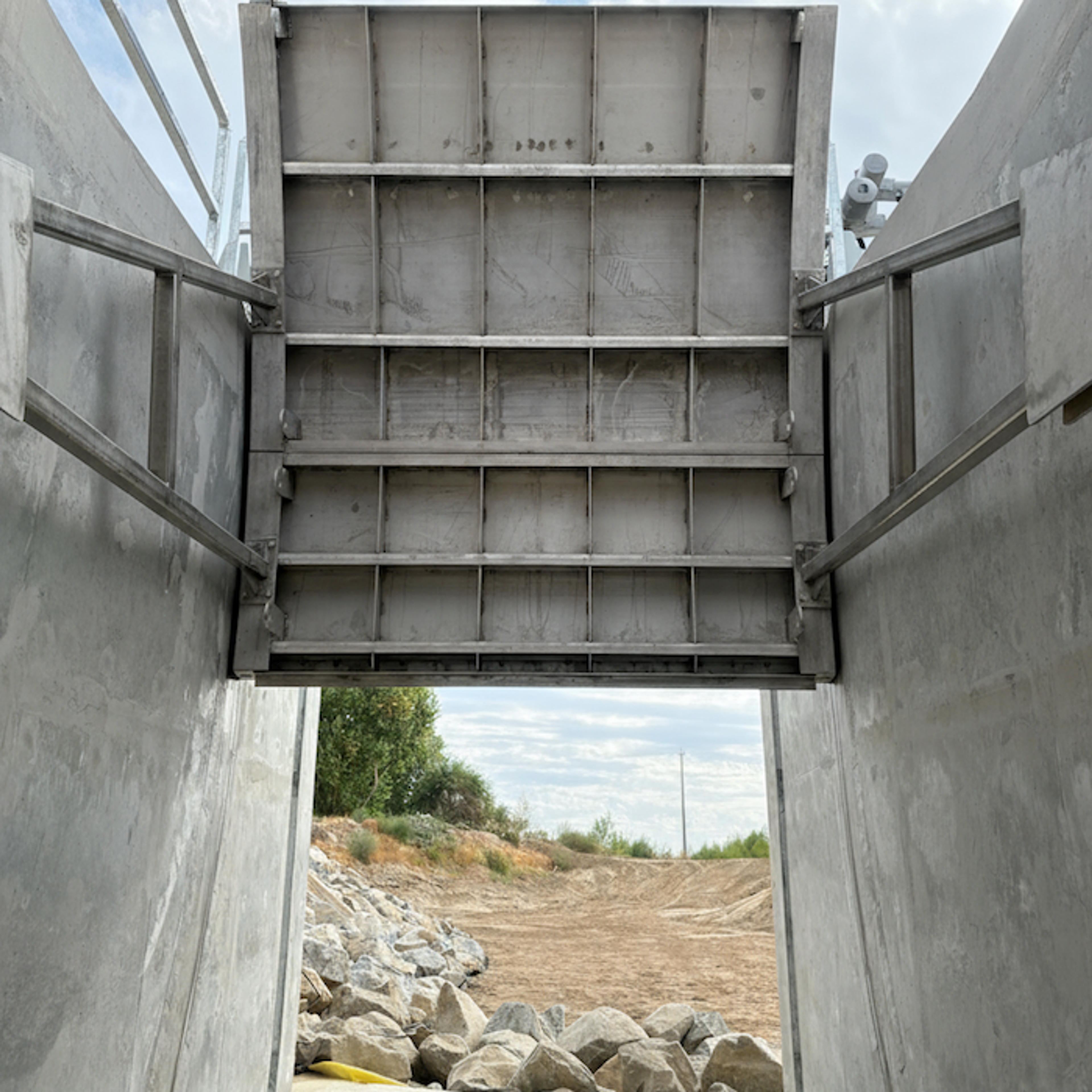 A large metal floodgate is partially open above a concrete channel, revealing rocks, dry ground, grass, and trees outside under a cloudy sky.