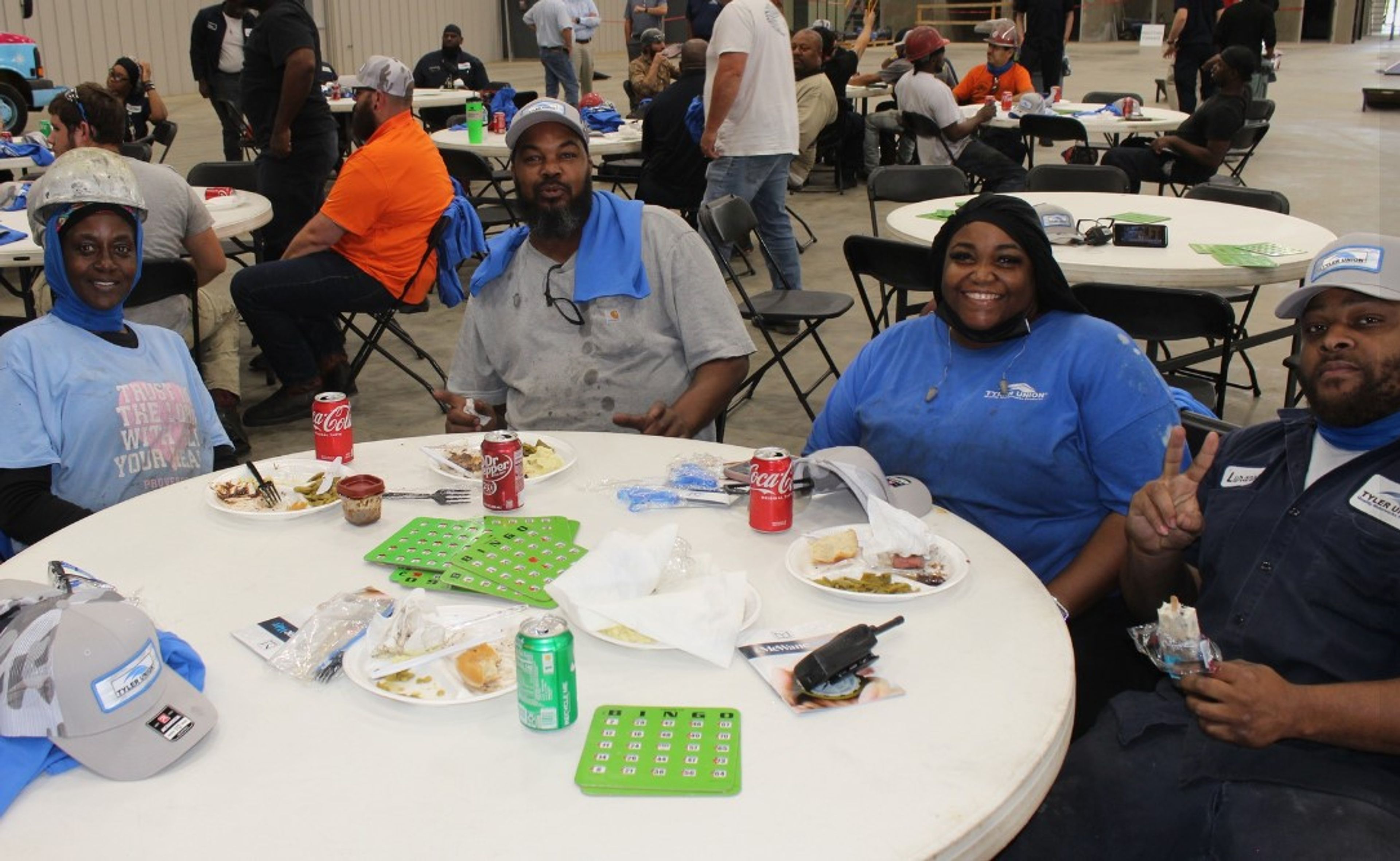 Four people sit at a round table enjoying a meal, smiling at the camera. Bingo cards and food are on the table. Others are eating and interacting at tables in the background. Indoor setting with casual, work-like attire.