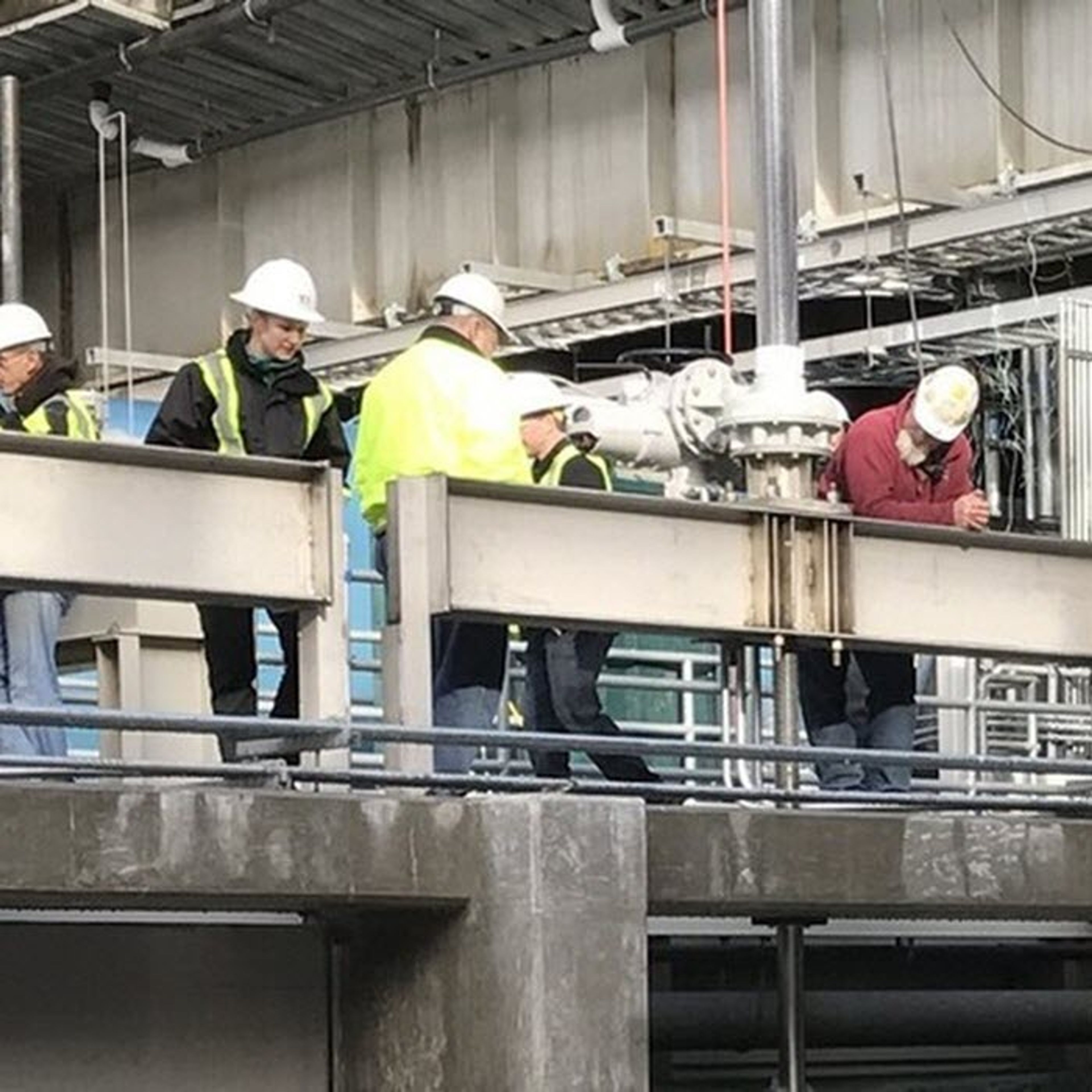 Construction workers in safety vests and helmets stand on a platform at an industrial site, inspecting machinery and metal structures under a partially finished roof.