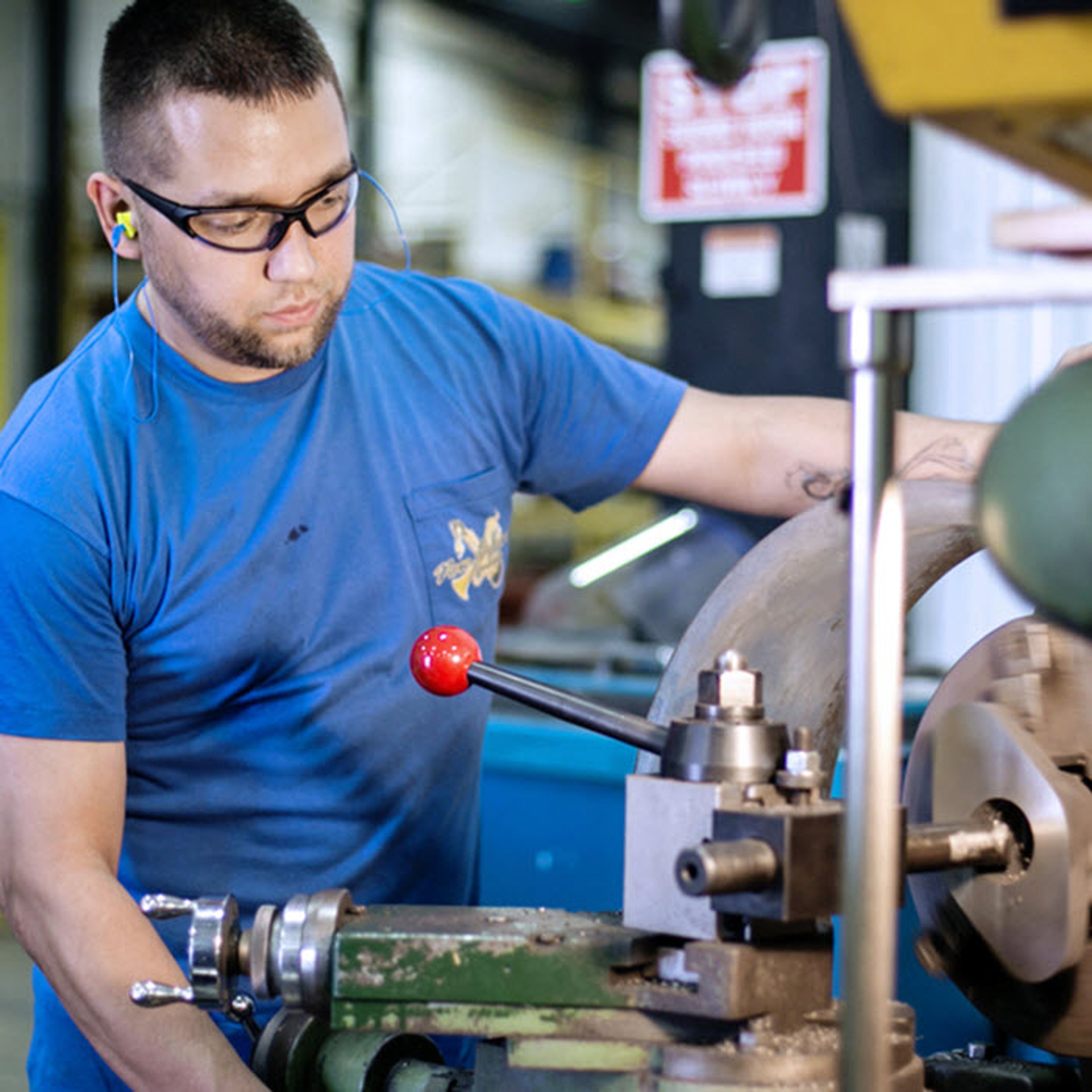 A man wearing safety glasses and earplugs operates a large industrial machine in a workshop, turning a metal object.
