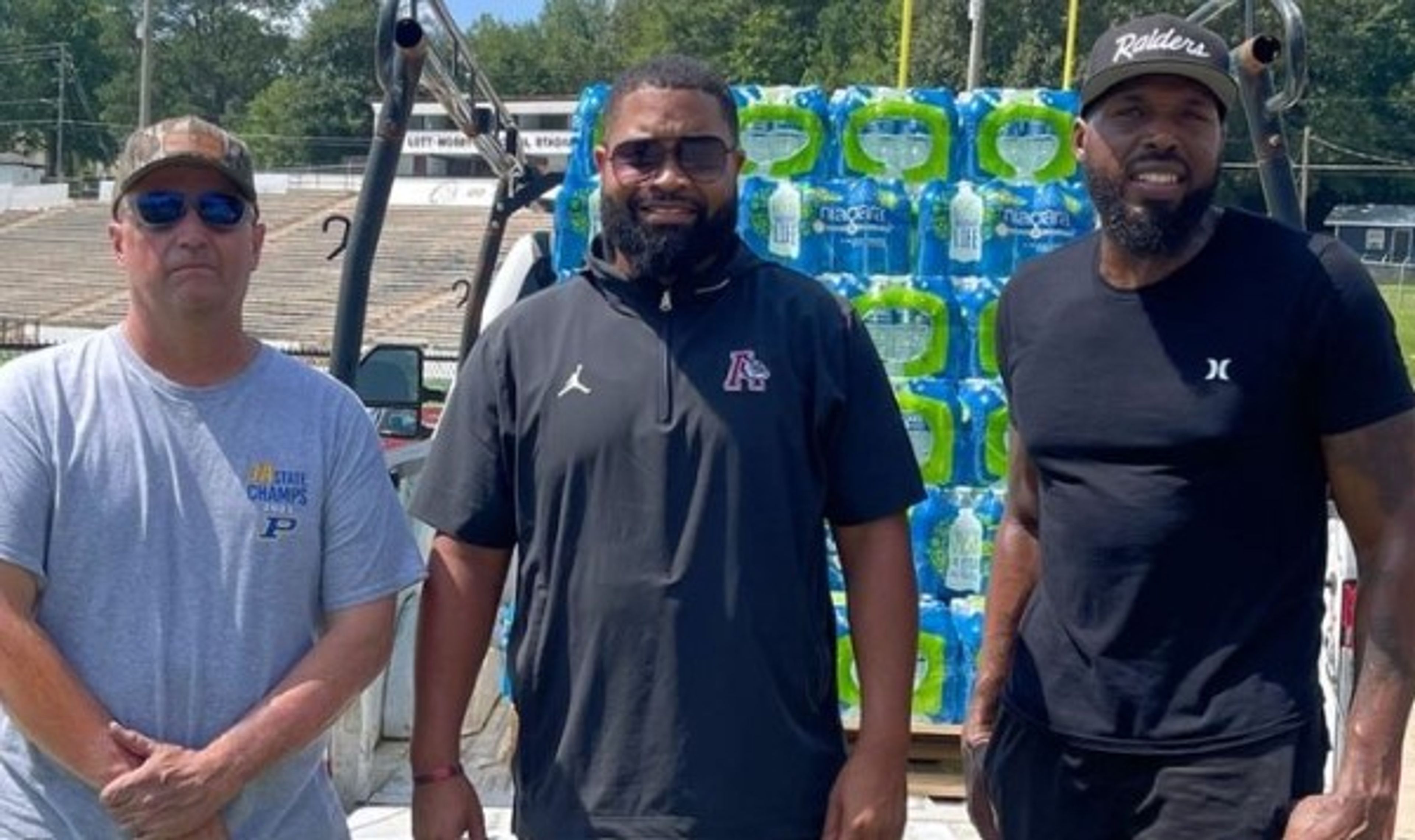 Three men stand in front of a truck loaded with packs of bottled water. They are outdoors on a sunny day, near a sports field with bleachers in the background.