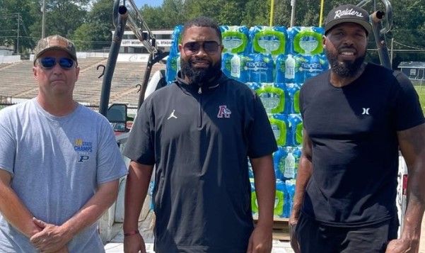 Three men stand in front of a truck loaded with packs of bottled water. They are outdoors on a sunny day, near a sports field with bleachers in the background.