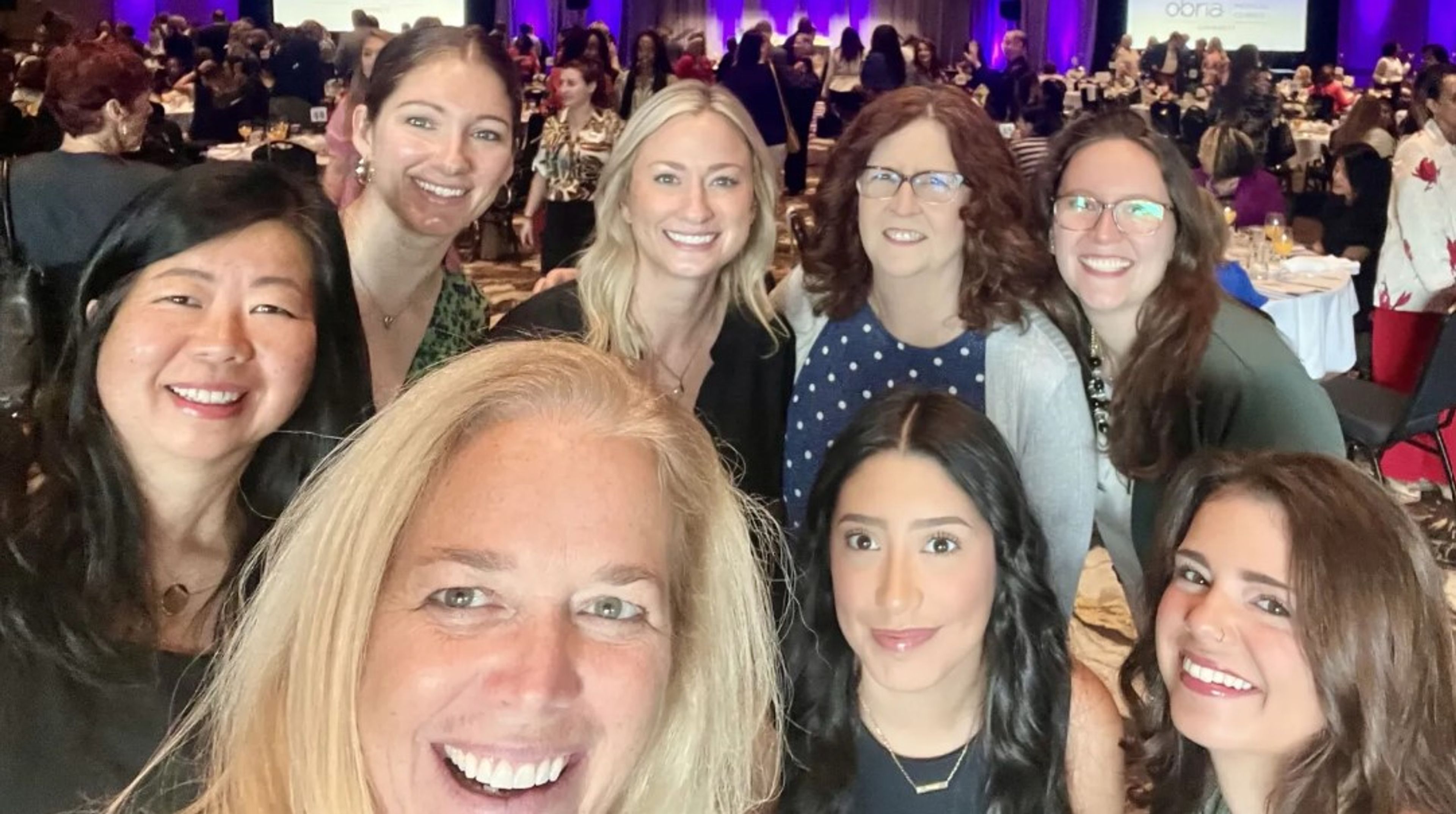 Eight women smile for a group selfie at an indoor event with round tables and other attendees visible in the background. The setting is well-lit, lively, and formal.