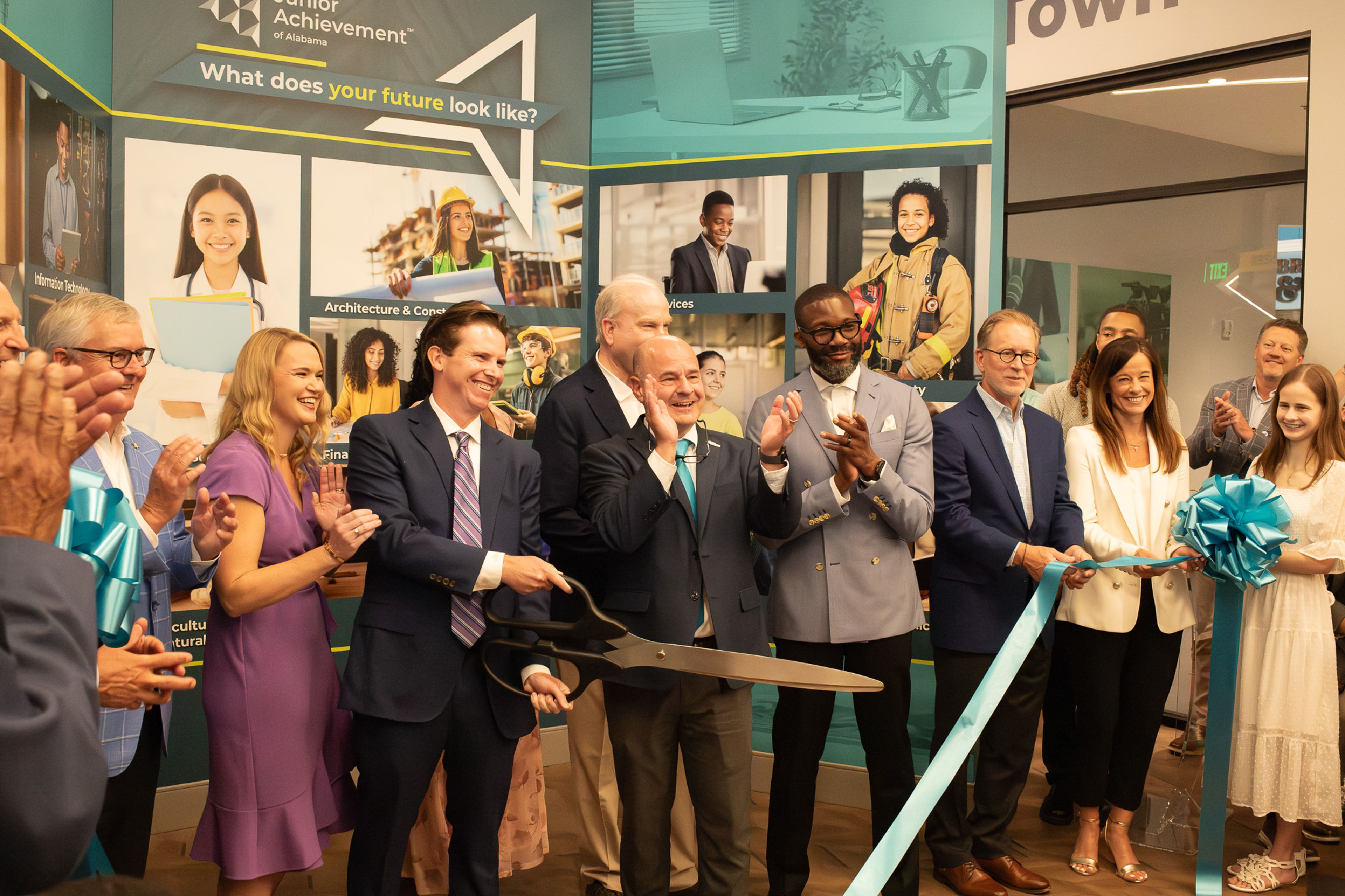 A group of people in business attire applauds as a man cuts a blue ribbon with large scissors at an indoor ribbon-cutting event. A display with career images is in the background.