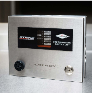 A stainless steel Amerex fire suppression control unit with a black display screen, labeled buttons, and connectors, sits on a metal surface.