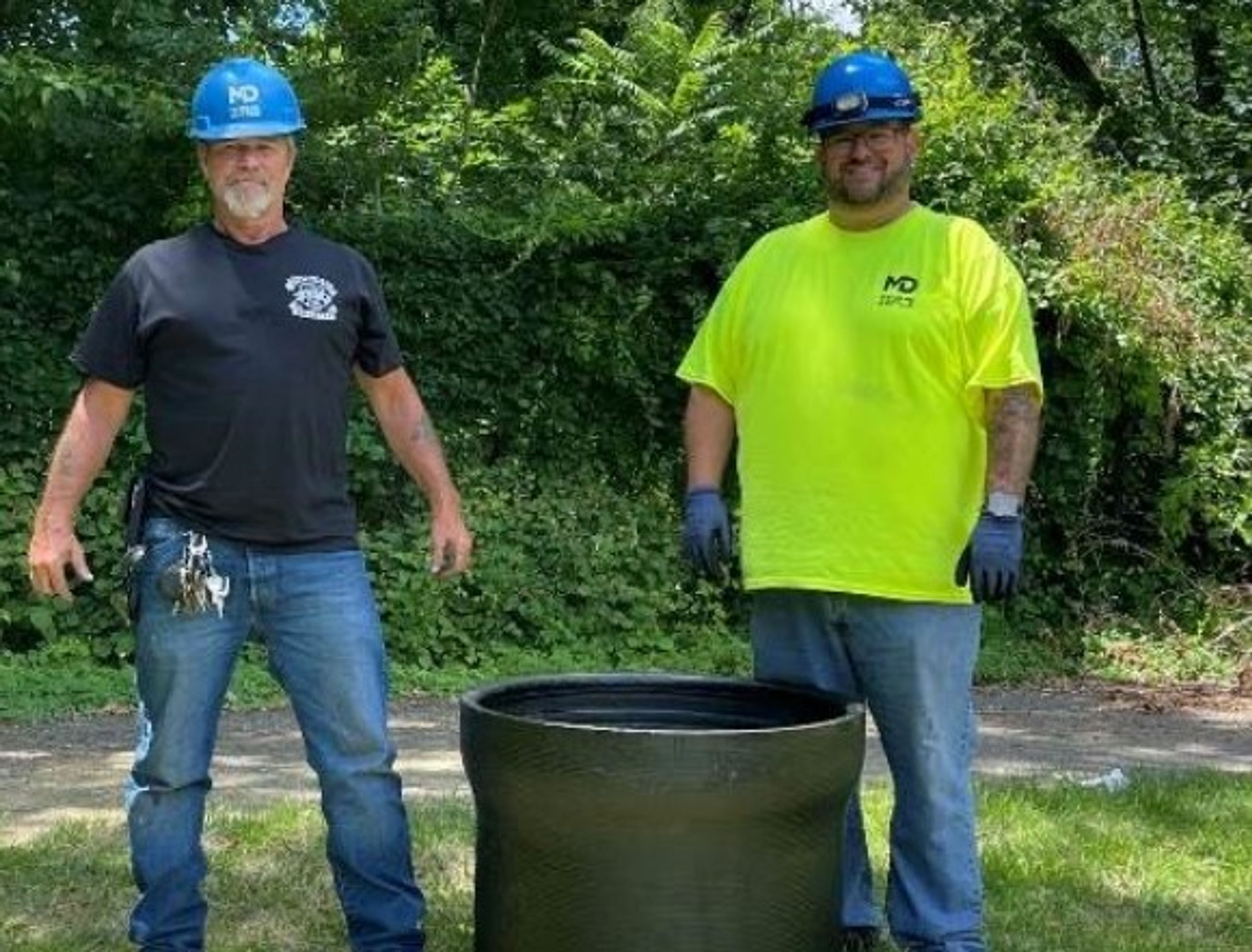Two workers wearing blue hard hats and gloves stand outdoors near a large black pipe. One wears a black shirt and jeans, and the other a bright yellow shirt and blue pants. Dense green foliage is in the background.