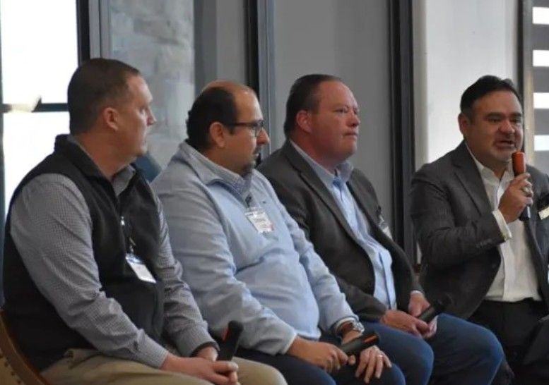 Four men sit in a row at a panel discussion. Three hold microphones in their laps while the man at the far right speaks into his microphone. They are dressed in business attire and seated indoors.